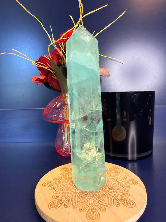 A bright green translucent fluorite crystal tower on a decorative wooden coaster in front of a red flower and black jar
