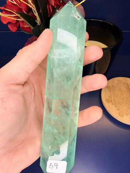 Hand holding a large translucent green fluorite tower in front of a red flower and black candle jar next to a wooden coaster