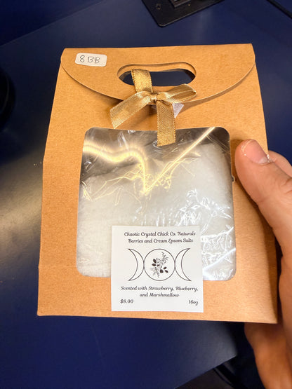 Hand holding white epsom salt in a brown paper container with a bow on a blue background