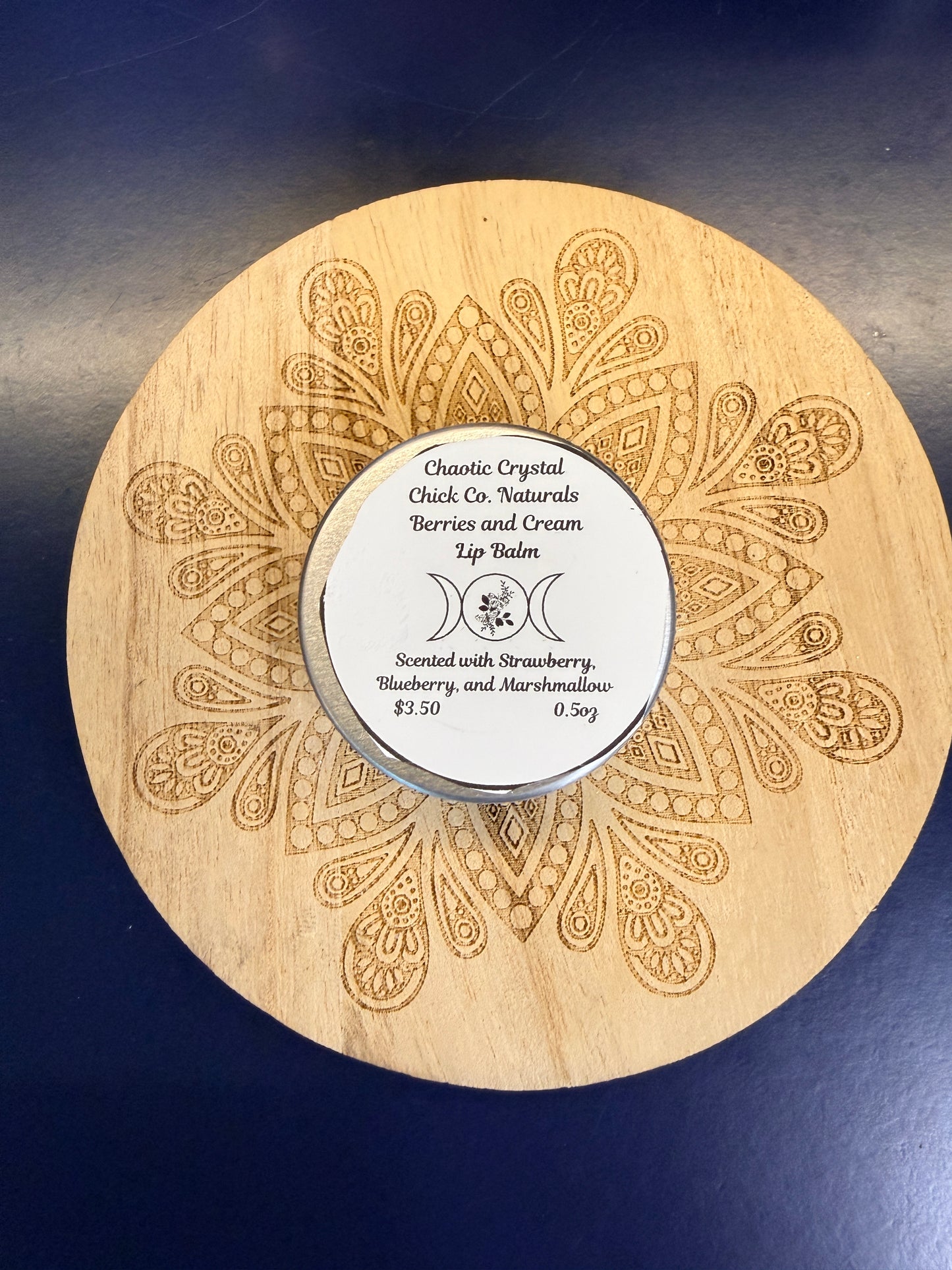 Wooden coaster with intricate patterns and a round metal lip balm on a dark blue background