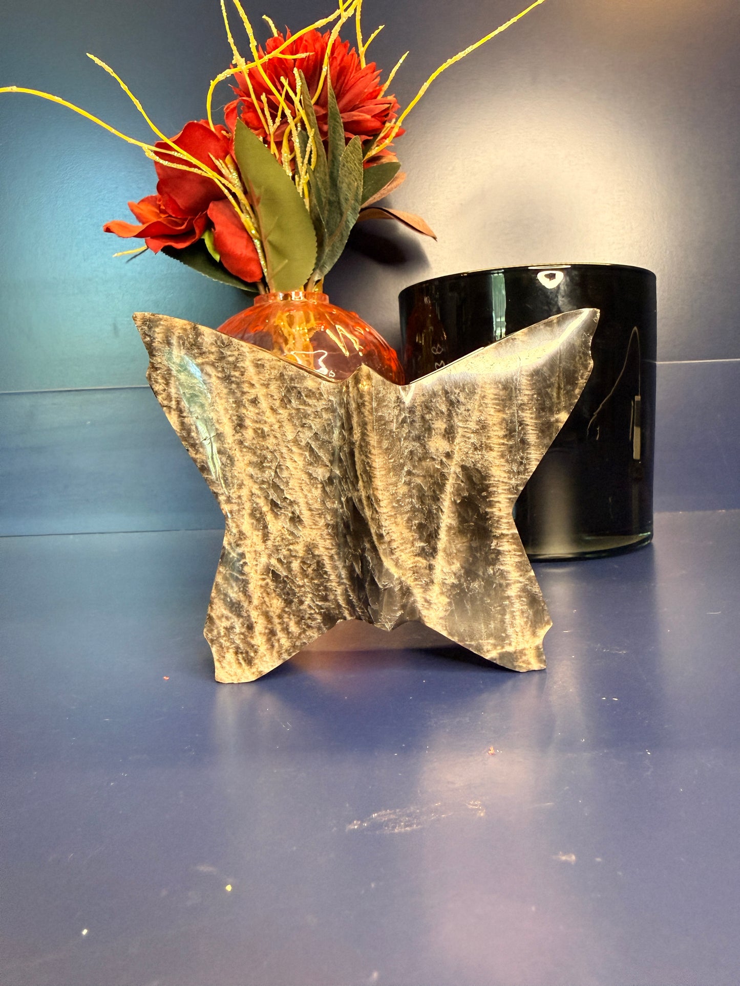 Large black moonstone butterfly in front of a pink mushroom vase and black jar on a blue background