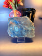A large rough blue apatite slab sitting in front of a mushroom vase and black jar