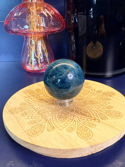 A blue apatite sphere on a clear stand sitting on a decorative wooden coaster in front of a pink mushroom and black jar