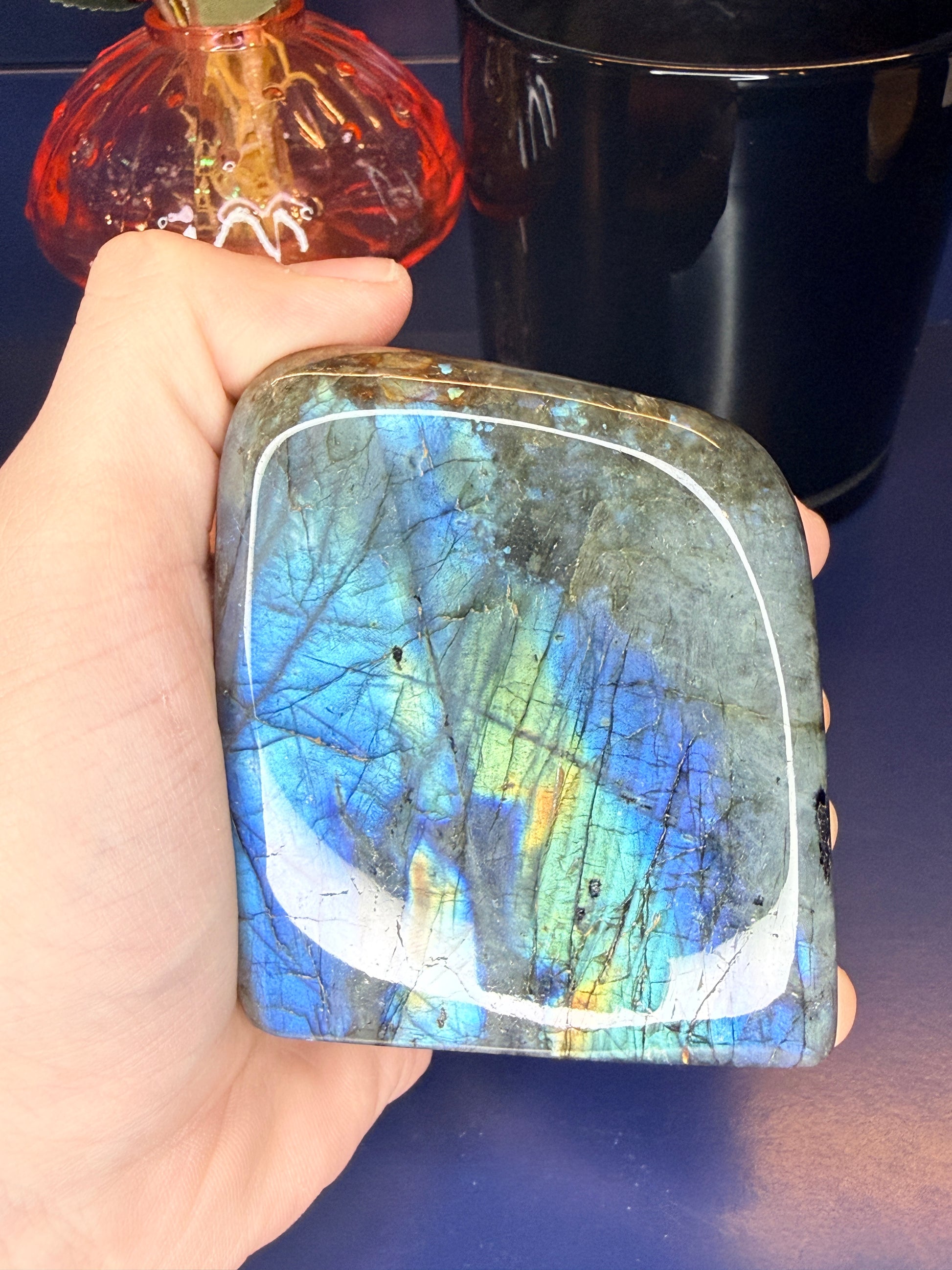 Hand holding a colorful blue flashing labradorite freeform in front of a mushroom vase and black jar