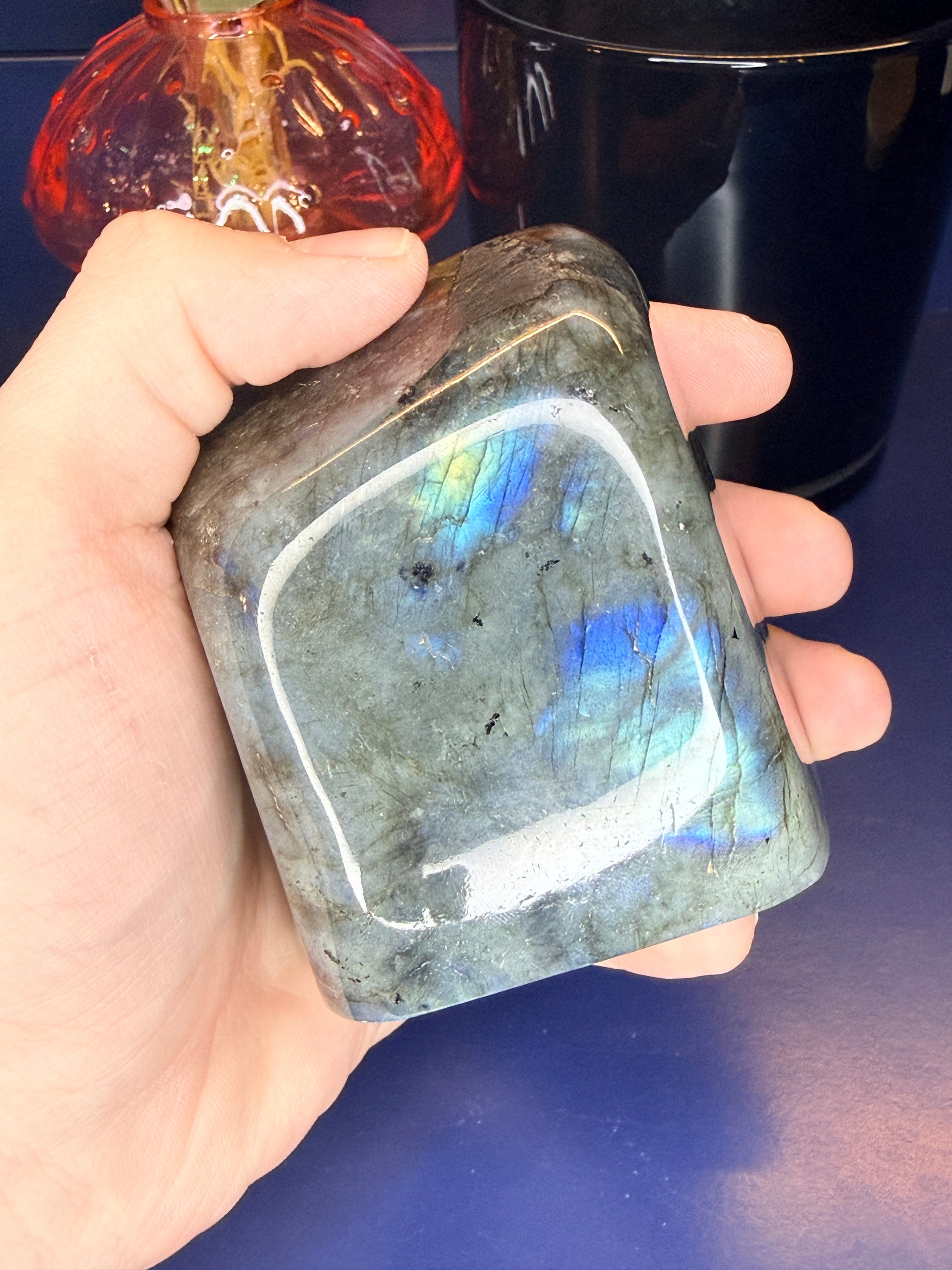Hand holding a colorful blue flashing labradorite freeform in front of a mushroom vase and black jar