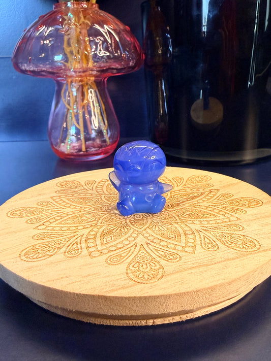 A blue opalite piplup crystal sitting on a decorative wooden coaster in front of a glass mushroom and black jar