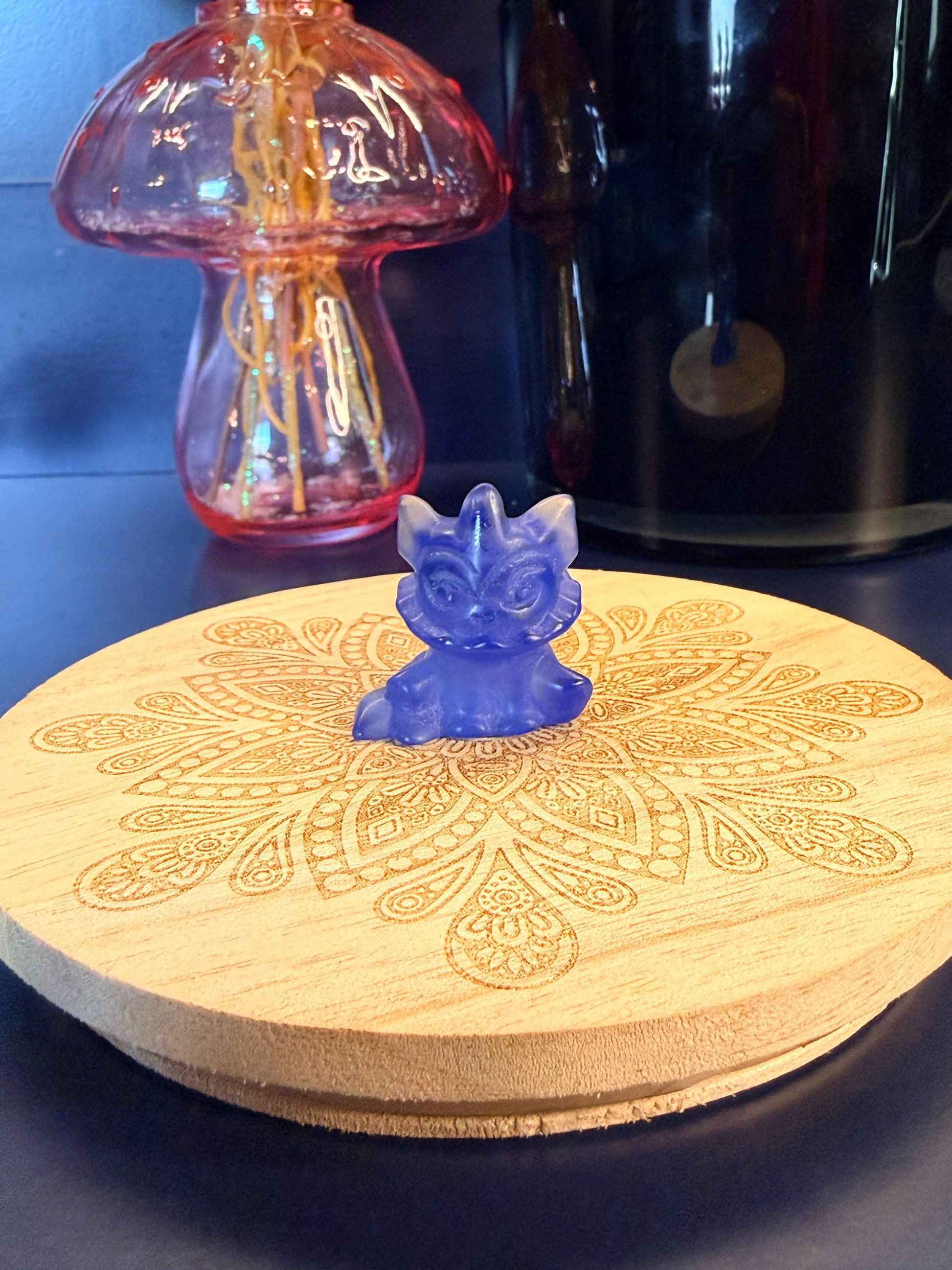 A blue opalite vaporeon crystal sitting on a decorative wooden coaster in front of a glass mushroom and black jar