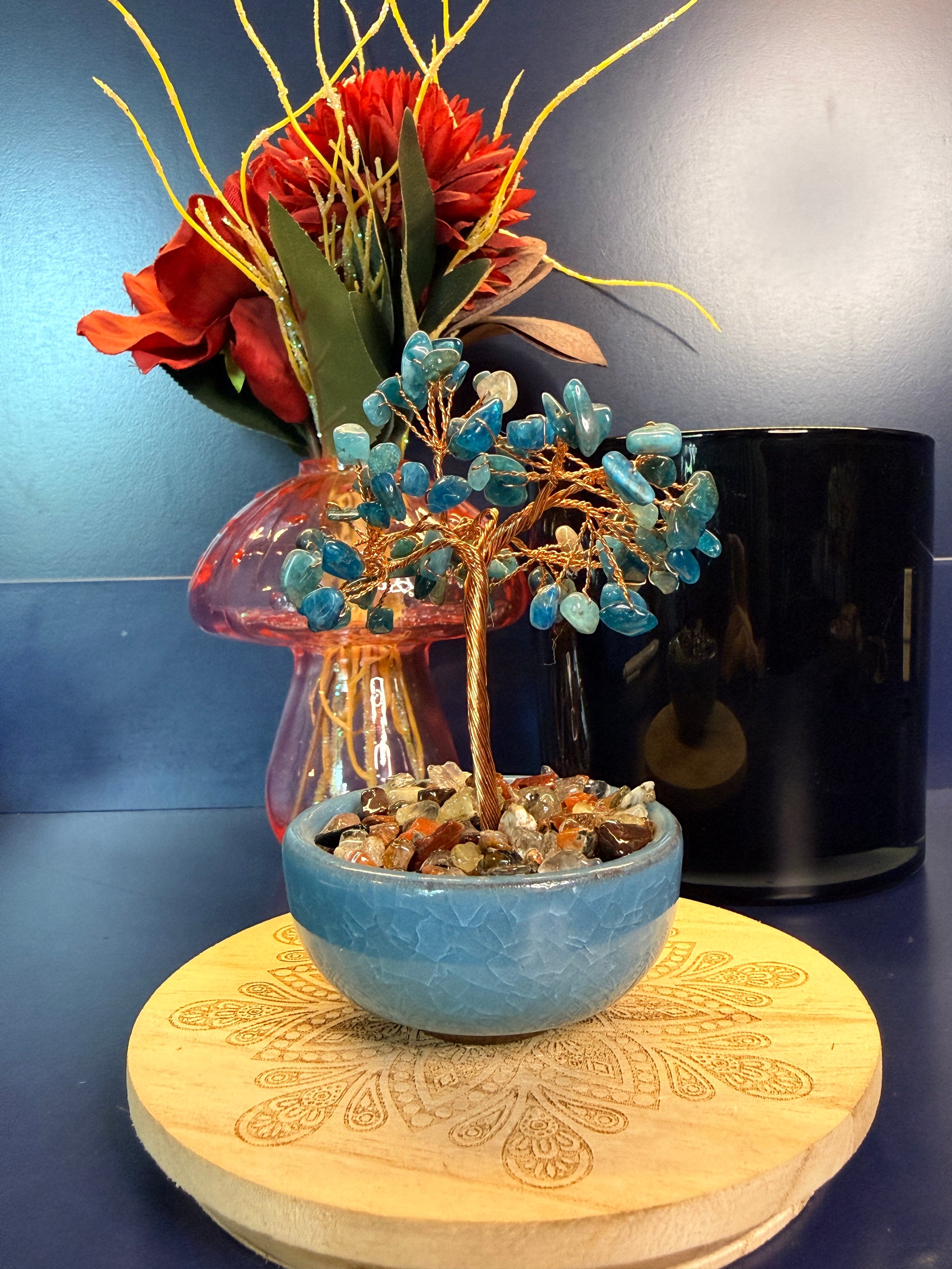 A blue crystal chip tree made of copper wire in a blue bowl on a wooden coaster in front of a pink mushroom and black jar