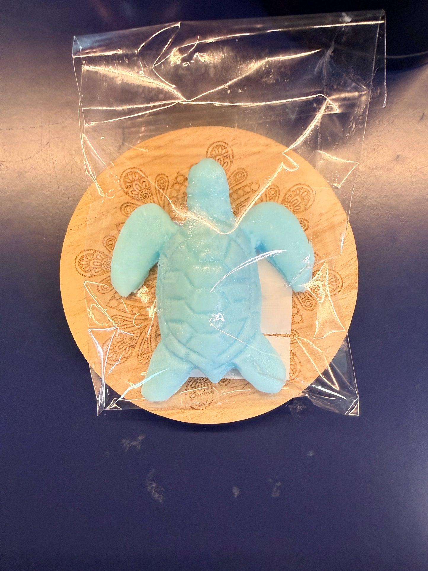 Blue turtle soap bar on a wooden coaster, wrapped in clear plastic on a dark surface.