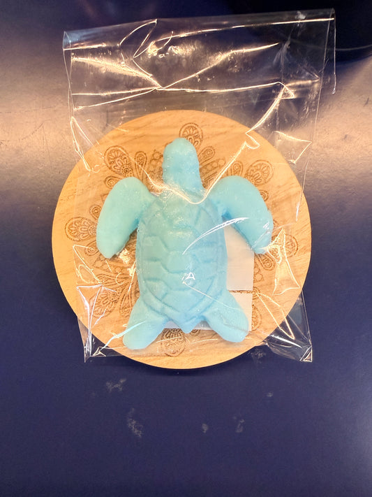 Blue turtle soap bar on a wooden coaster, wrapped in clear plastic on a dark surface.