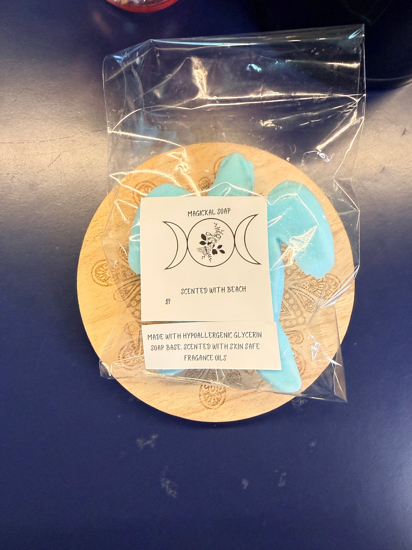 Blue turtle soap bar with two labels on a wooden coaster, wrapped in clear plastic on a dark surface.