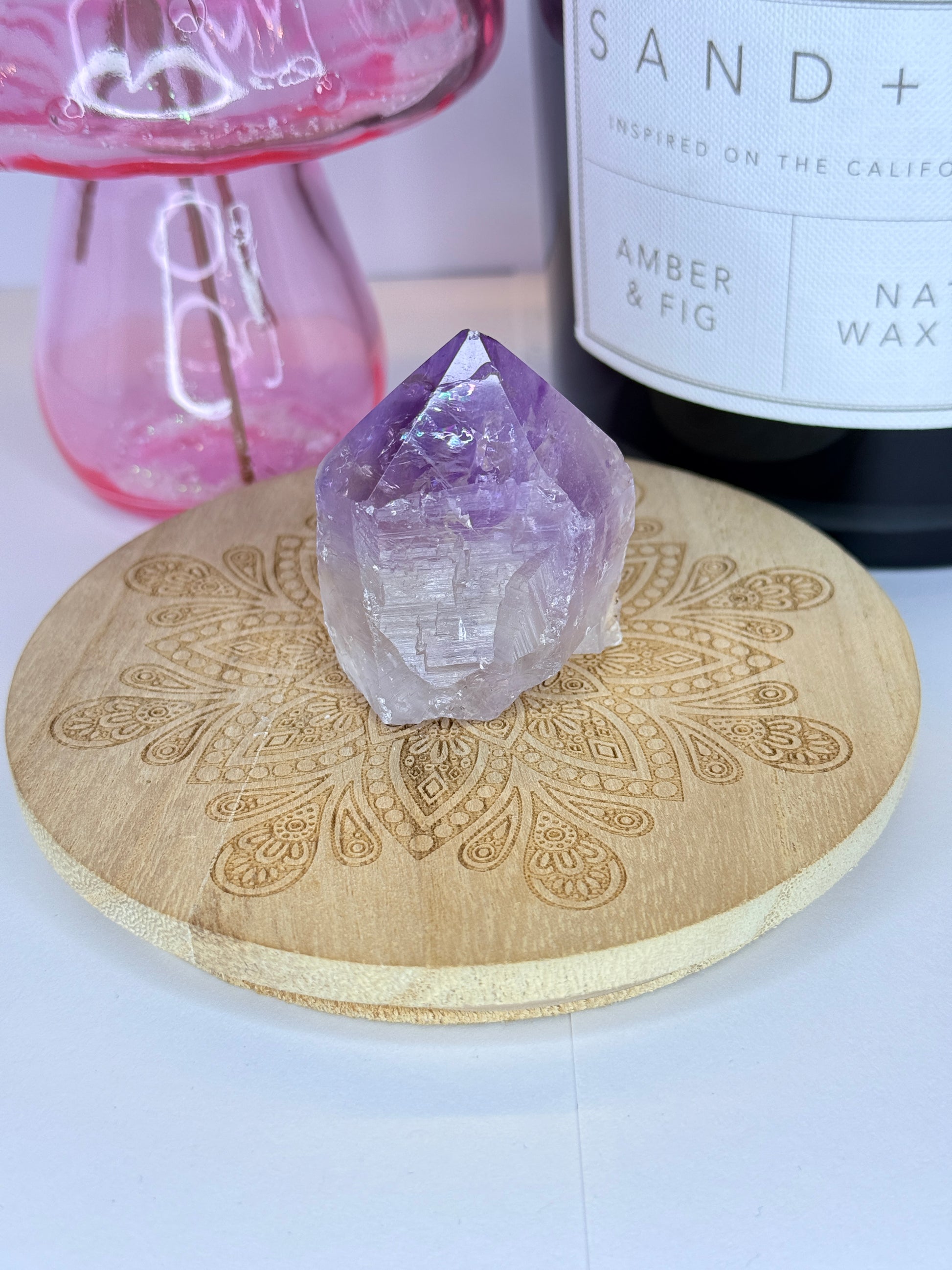Purple and white amethyst raw tower resting on a wooden coaster in front of a black candle jar and pink vase
