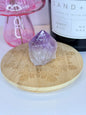 Purple and white amethyst raw tower resting on a wooden coaster in front of a black candle jar and pink vase