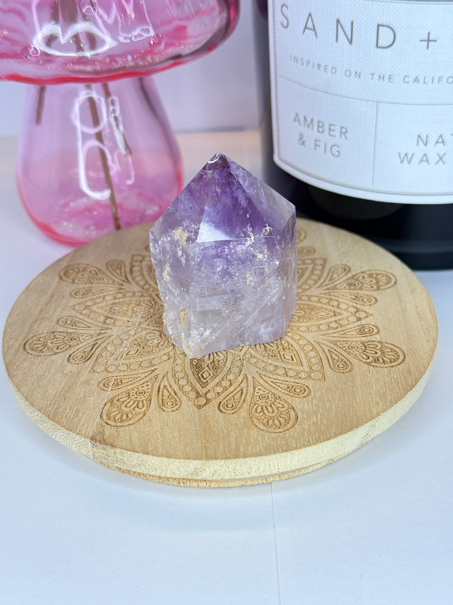 Purple and white amethyst raw tower resting on a wooden coaster in front of a black candle jar and pink vase