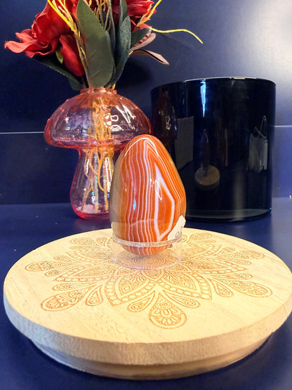 A red and white crystal egg sitting on a decorative wooden coaster in front of a glass mushroom and black jar