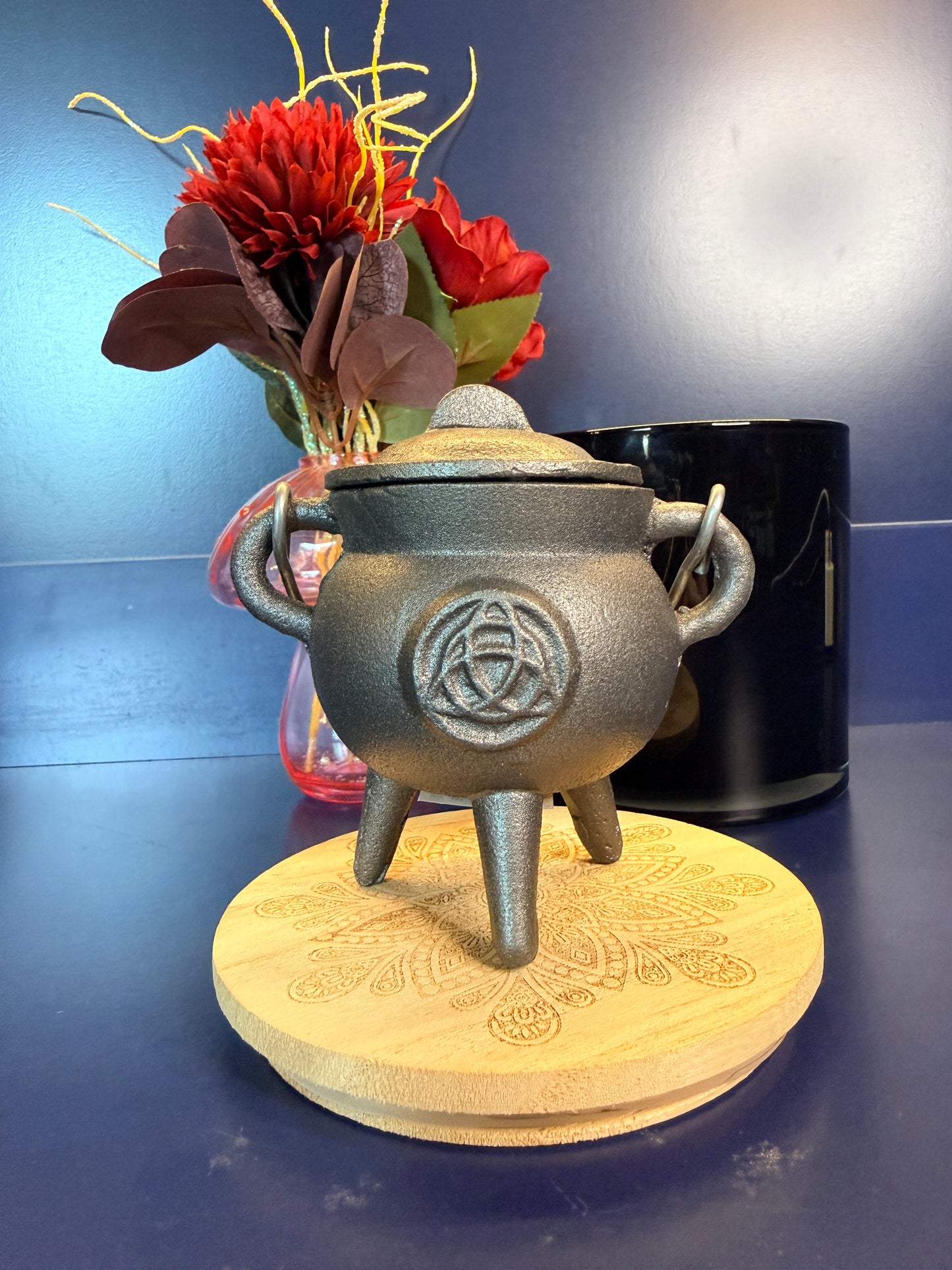 A cast iron cauldron with a celtic knot design sitting on a decorative wooden coaster in front of a red flower and black jar