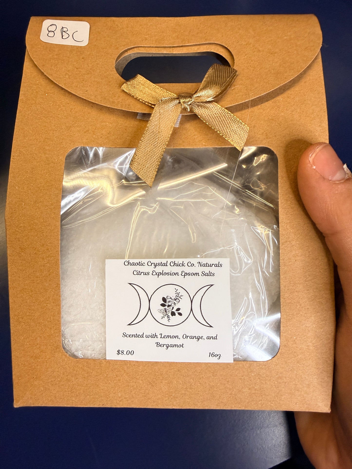Hand holding white epsom salt in a brown paper container with a bow on a blue background