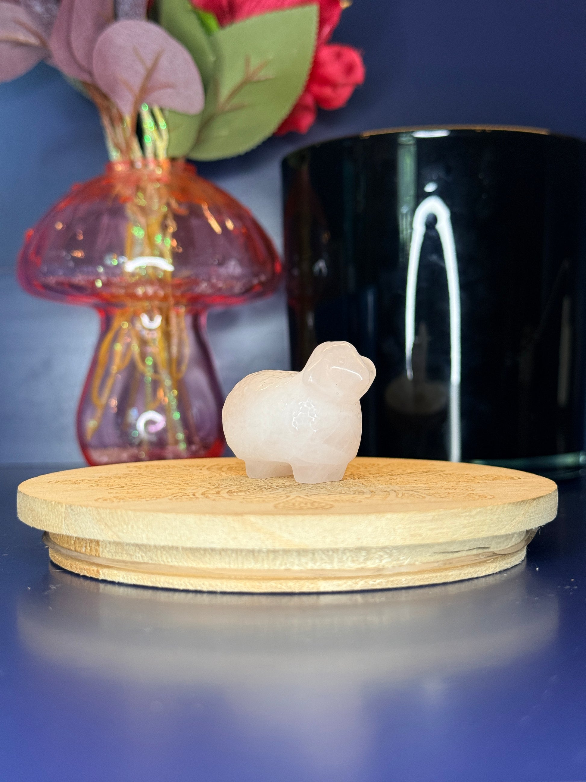 Clear quartz sheep carving in front of a pink vase and black jar on a wooden coaster against a blue background