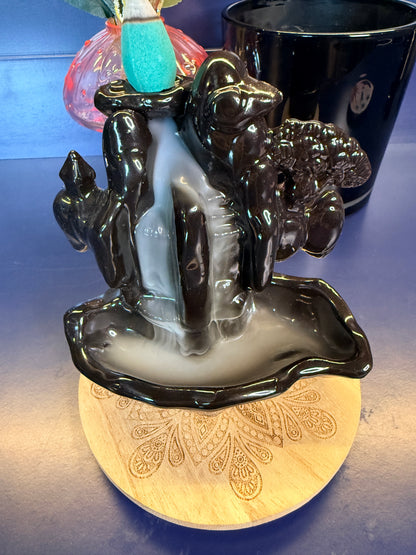 Black waterfall incense burner covered in white smoke with a blue incense cone on top sitting on a decorative wooden coaster.