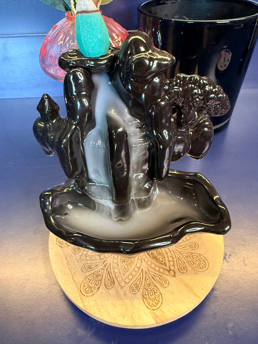 Black waterfall incense burner covered in white smoke with a blue incense cone on top sitting on a decorative wooden coaster.
