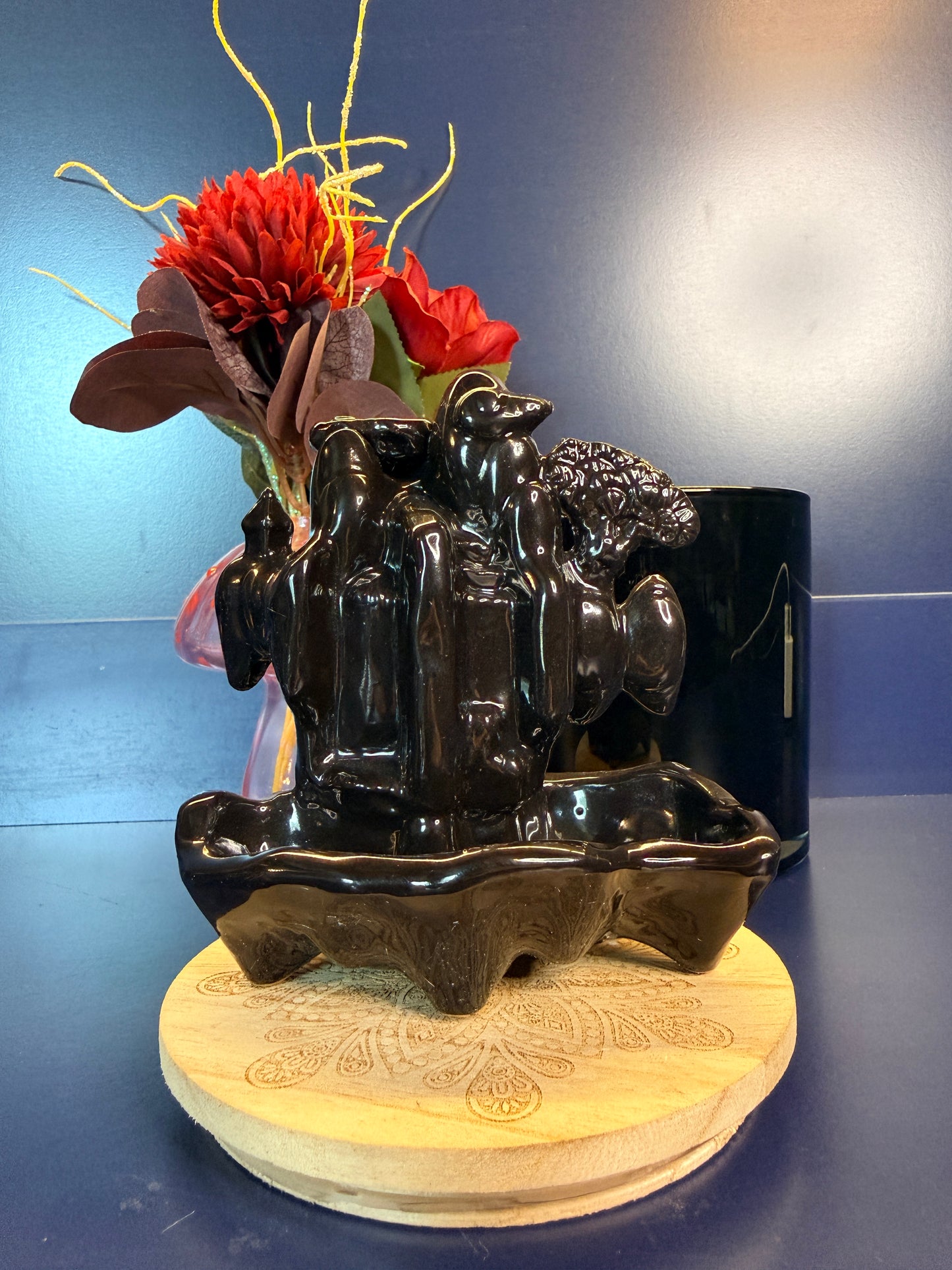 Black waterfall incense burner in front of  red flower sitting on a decorative wooden coaster against a blue background