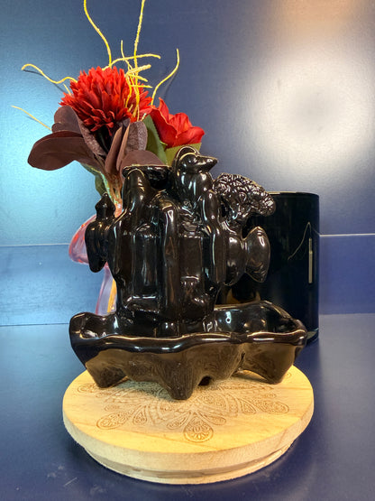 Black waterfall incense burner in front of  red flower sitting on a decorative wooden coaster against a blue background