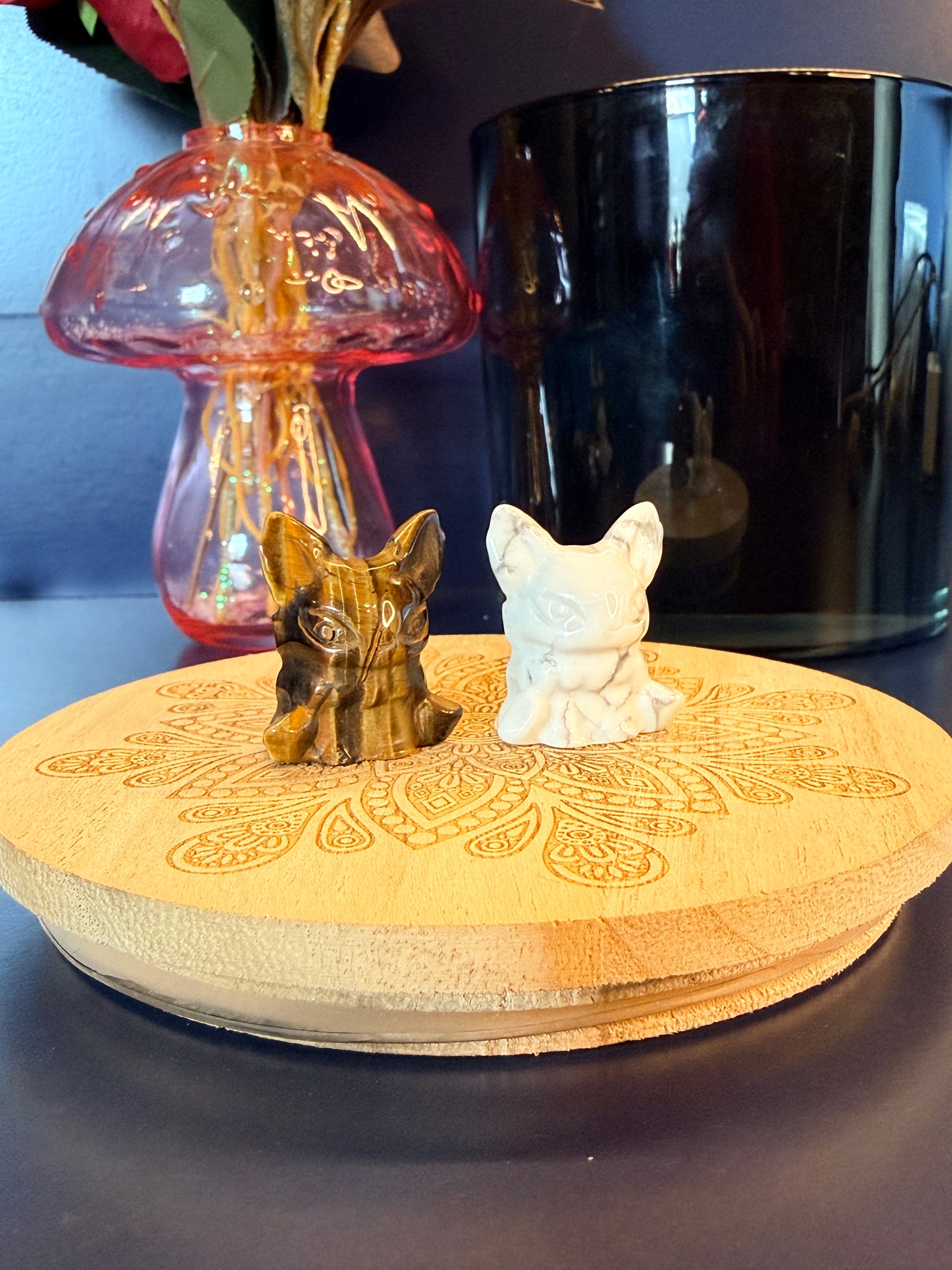 Two crystal aliens sitting on a decorative wooden coaster in front of a glass mushroom and black jar