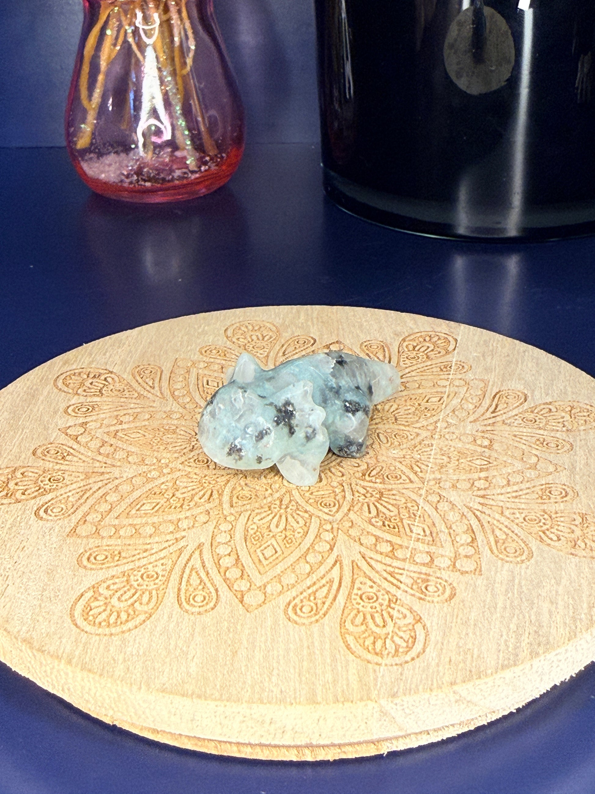 green and black kiwi jasper axolotl carving in front of a pink vase and black jar on a wooden coaster against a blue background