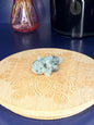 green and black kiwi jasper axolotl carving in front of a pink vase and black jar on a wooden coaster against a blue background