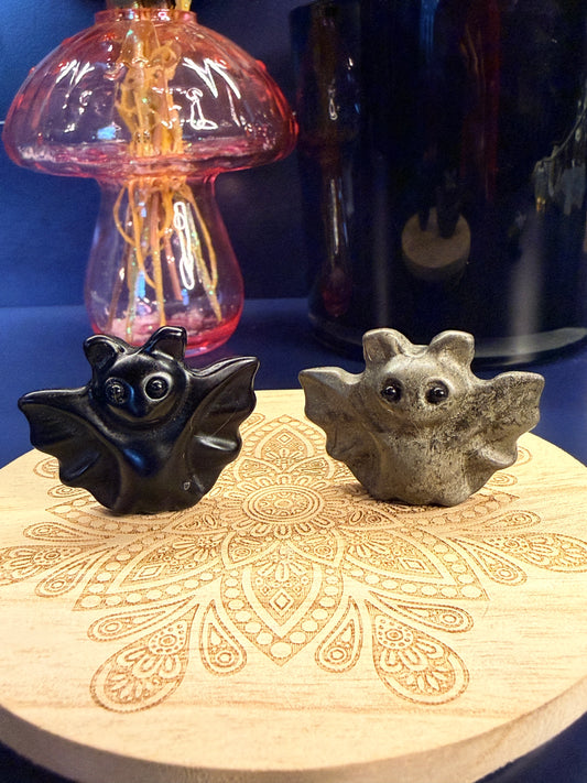 Two crystal bats sitting on a decorative wooden coaster in front of a glass mushroom and black jar