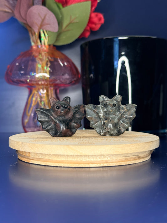 black and grey crystal bat carvings in front of a pink vase and black jar on a wooden coaster against a blue background 