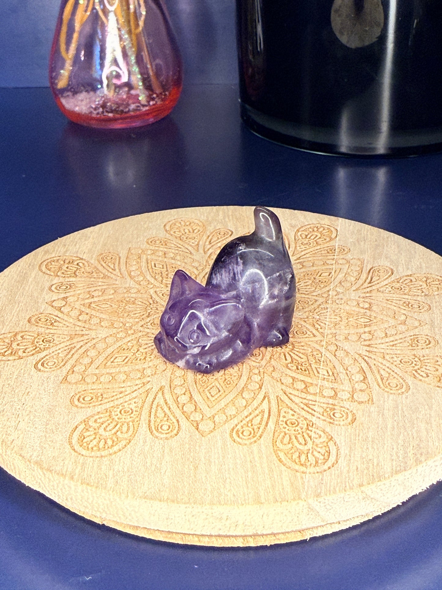 Purple amethyst cat carving in front of a pink vase and black jar on a wooden coaster against a blue background