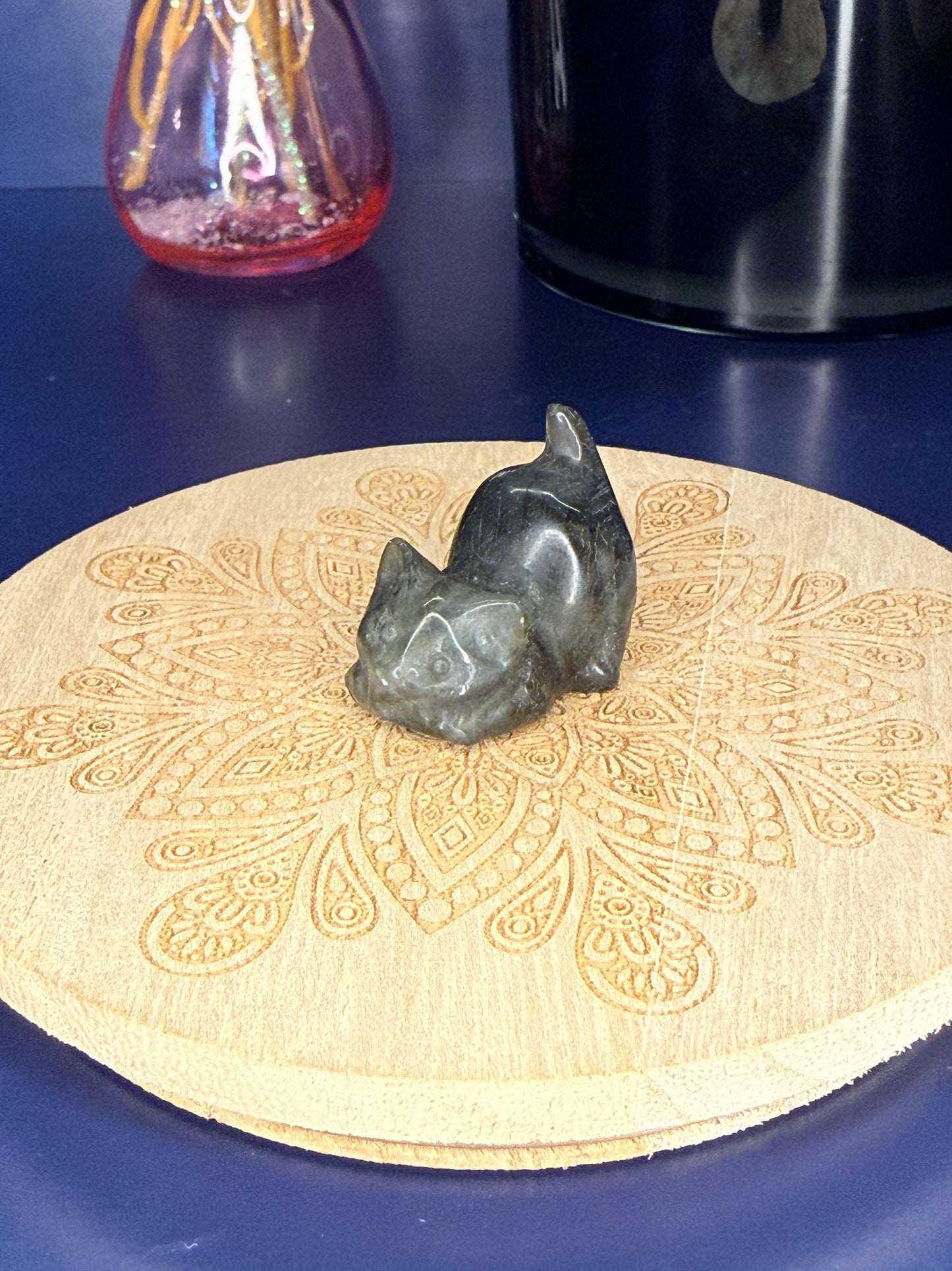 Grey labradorite cat carving in front of a pink vase and black jar on a wooden coaster against a blue background