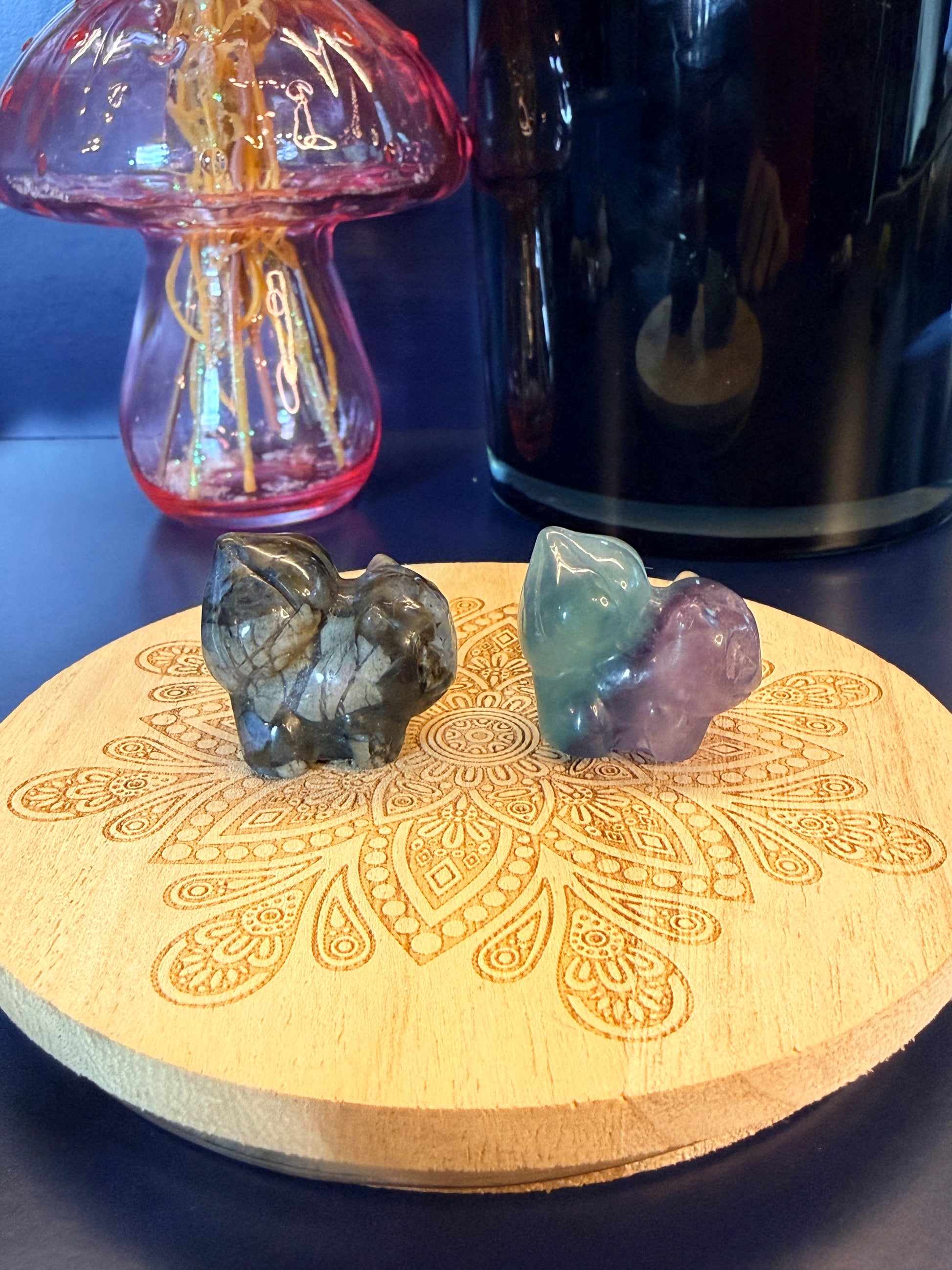 Two crystal shapes, one que sera and one fluorite, carved to resemble a bulbasaur-inspired creature sitting on a decorative wooden coaster in front of a black candle vessel and glass mushroom against a blue background