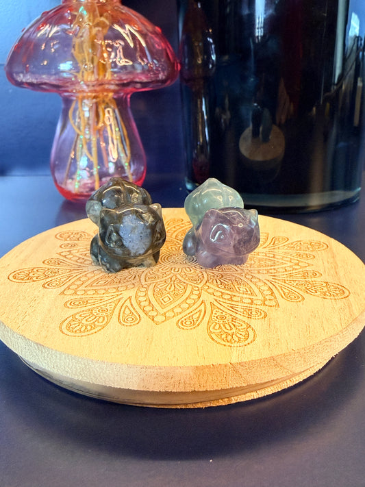 Two crystal shapes, one que sera and one fluorite, carved to resemble a bulbasaur-inspired creature sitting on a decorative wooden coaster in front of a black candle vessel and glass mushroom against a blue background