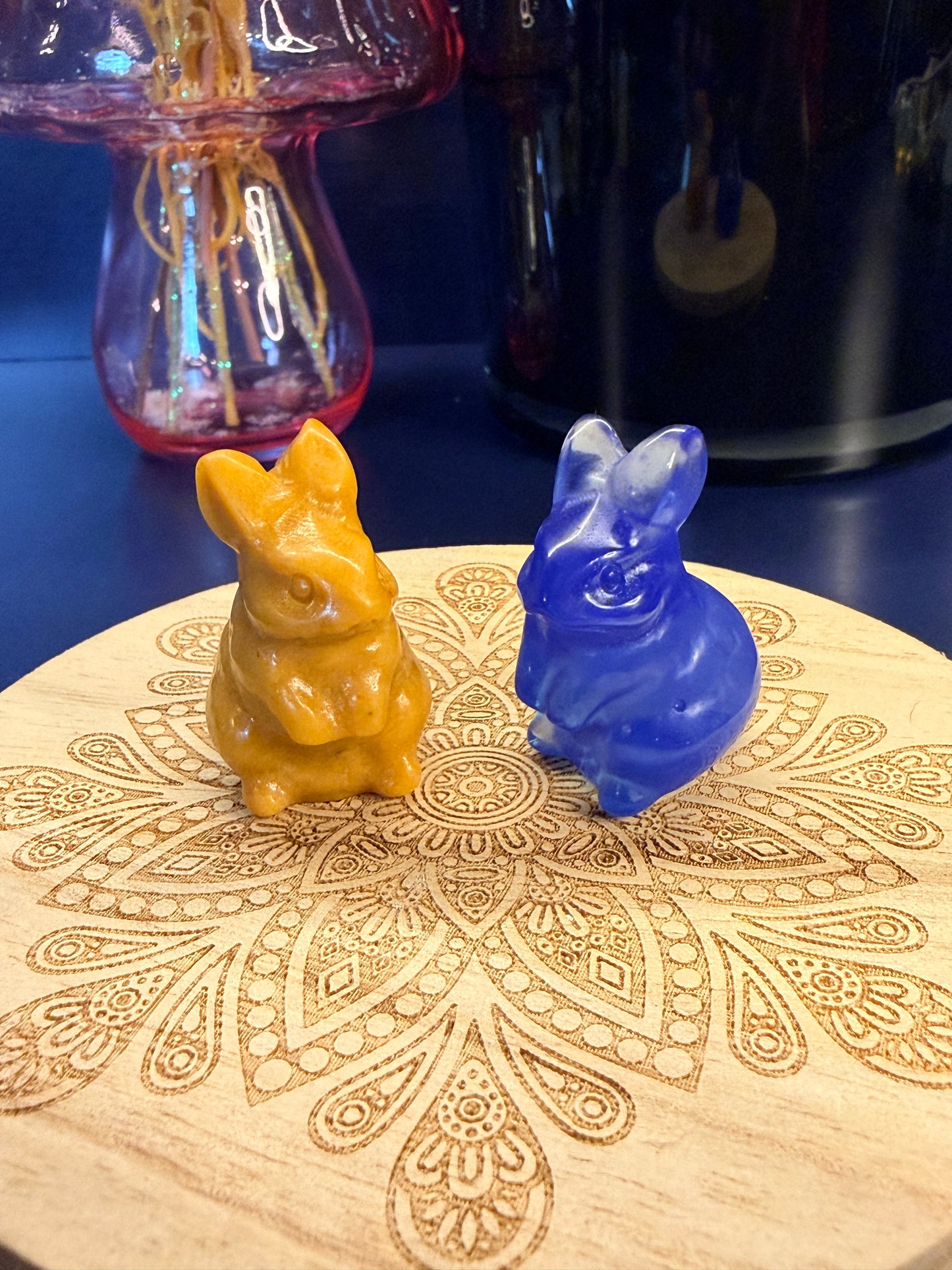 Two crystal bunnies sitting on a decorative wooden coaster in front of a glass mushroom and black jar