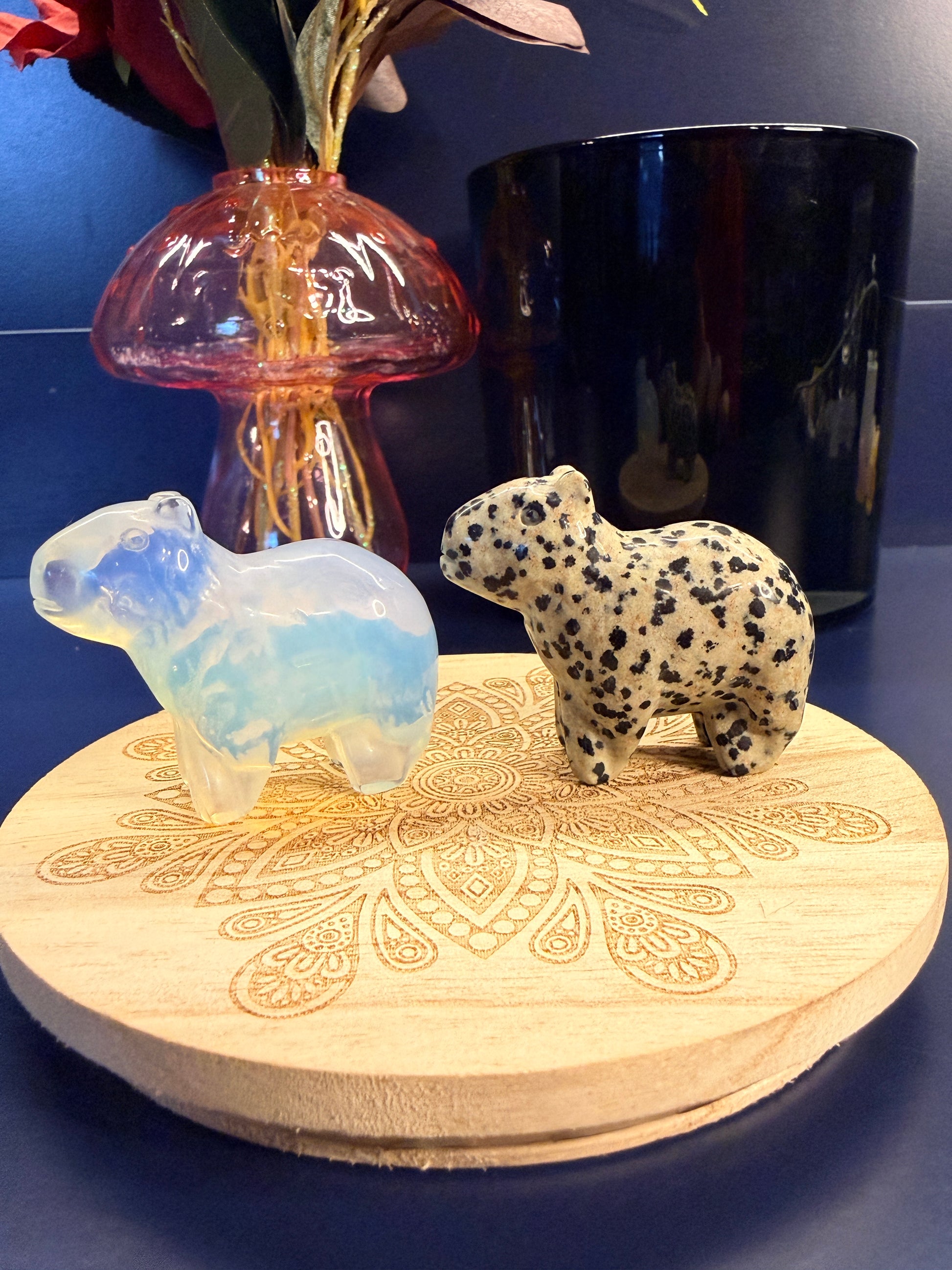 Two crystal Capybaras sitting on a decorative wooden coaster in front of a glass mushroom and black jar