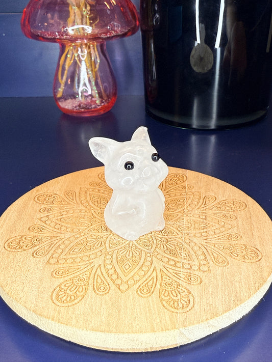 White Clear Quartz pig carving in front of a pink vase and black jar on a wooden coaster against a blue background