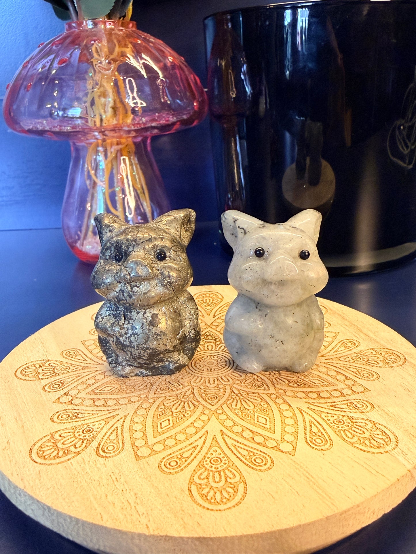 Two crystal cartoon pigs sitting on a decorative wooden coaster in front of a glass mushroom and black jar