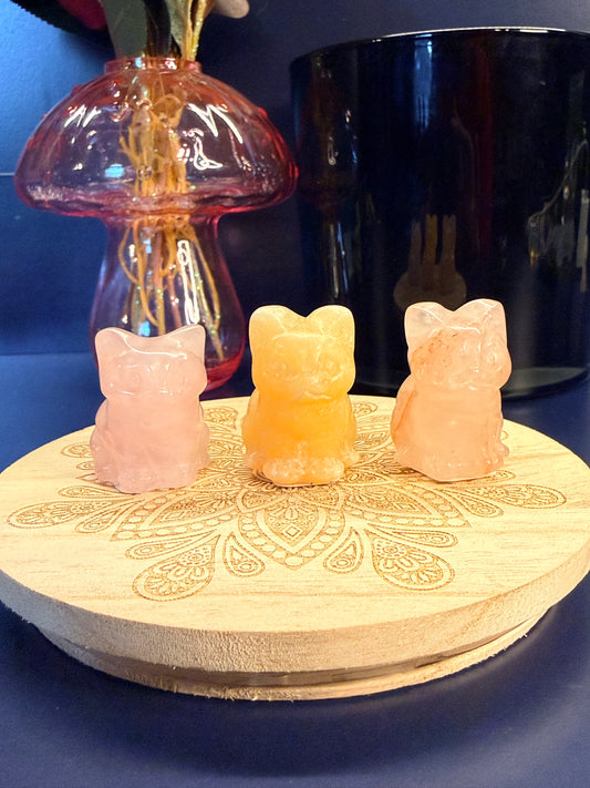 Three crystal cats sitting on a decorative wooden coaster in front of a glass mushroom and black jar