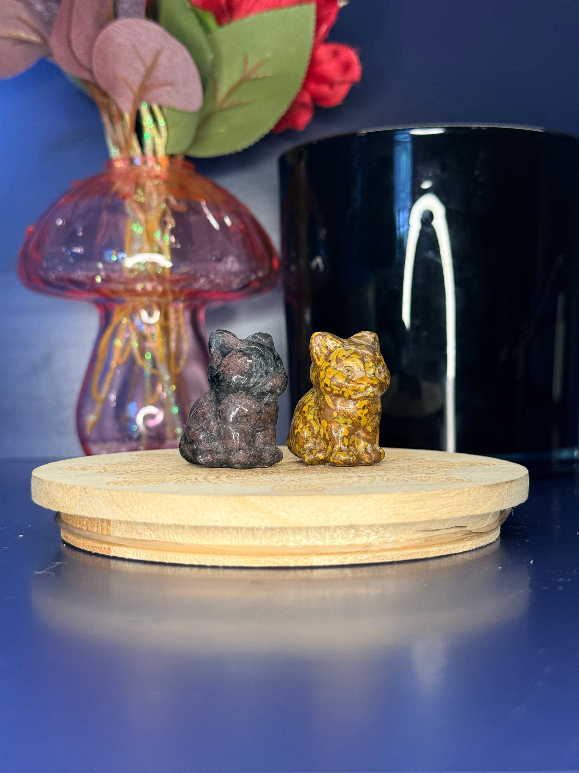 Black and yellow crystal cat carvings in front of a pink vase and black jar on a wooden coaster against a blue background