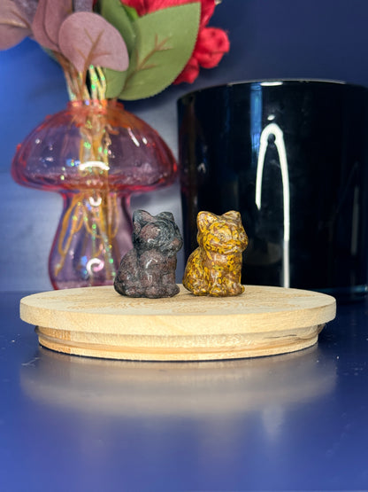 Black and yellow crystal cat carvings in front of a pink vase and black jar on a wooden coaster against a blue background