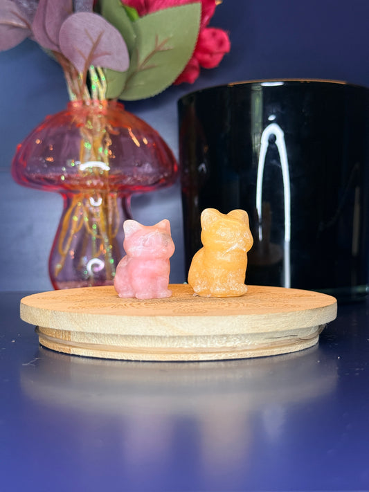 Pink and orange crystal cat carvings in front of a pink vase and black jar on a wooden coaster against a blue background