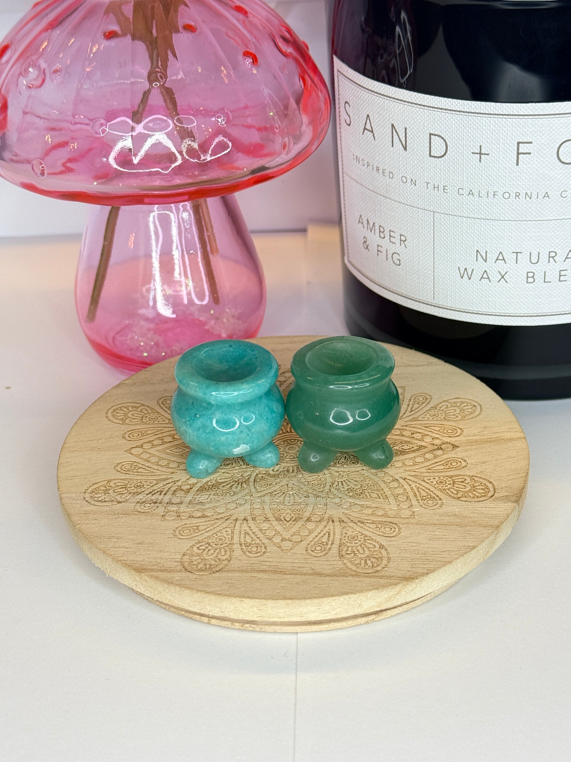 Two blue and green crystal cauldron carvings resting on a wooden coaster in front of a black jar and pink vase