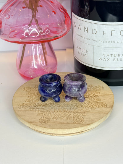 Two blue and purple crystal cauldron carvings resting on a wooden coaster in front of a black jar and pink vase