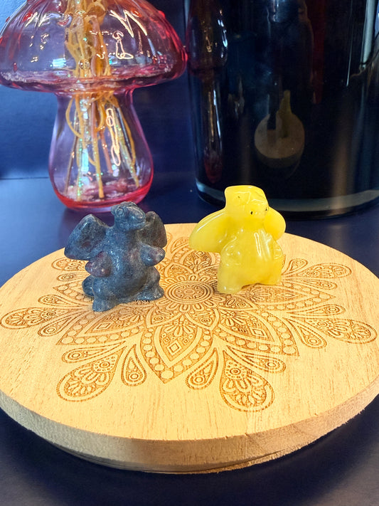 Two crystals, one blue calcite and one lemon calcite, carved into charizard-inspired creatures sitting on a decorative wooden coaster in front of a black candle vessel and a pink glass mushroom against a blue background