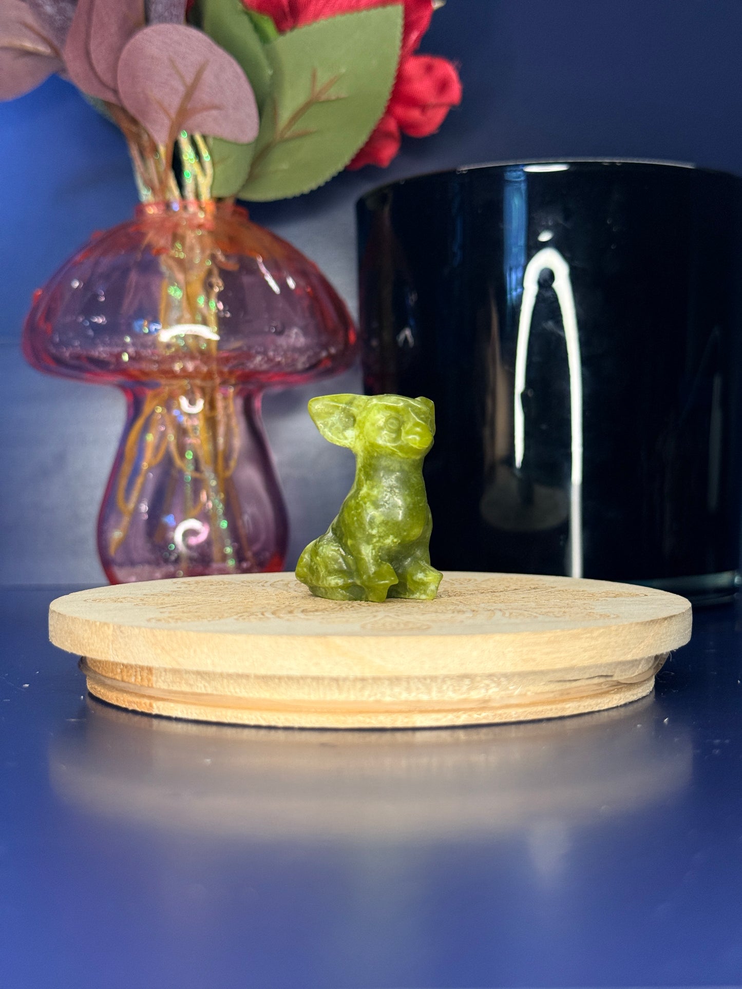 Green serpentine chihuahua carving in front of a pink vase and black jar on a wooden coaster against a blue background