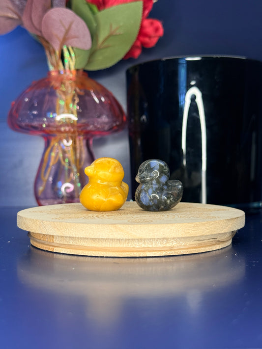 Orange and blue crystal duck carvings in front of a pink vase and black jar on a wooden coaster against a blue background