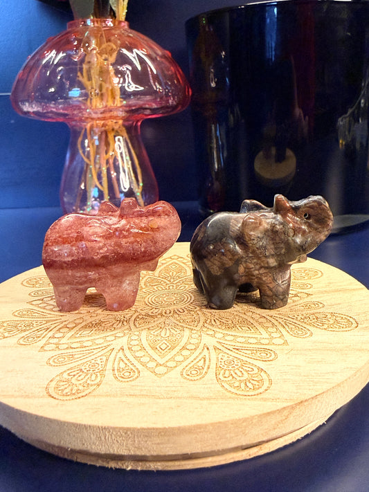 Two crystal elephants sitting on a decorative wooden coaster in front of a glass mushroom and black jar