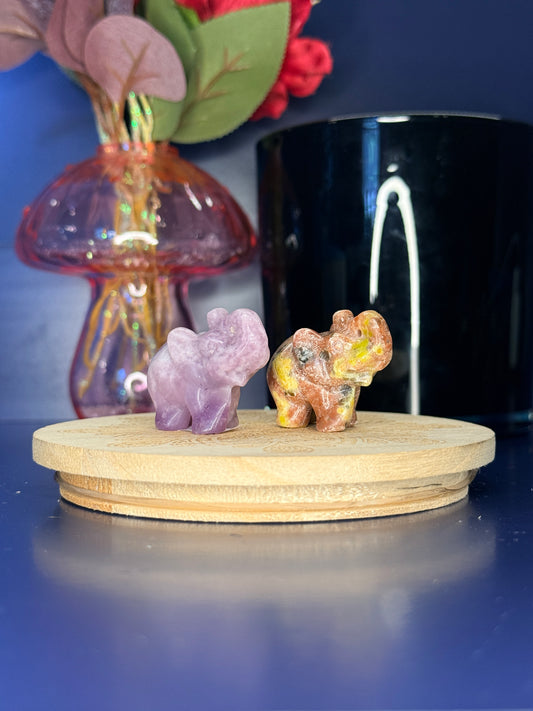 multicolored crystal elephant carvings in front of a pink vase and black jar on a wooden coaster against a blue background